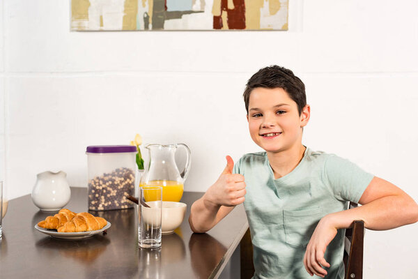 smiling boy sitting at table during breakfast and showing thumb up