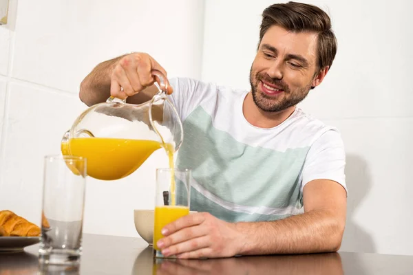 キッチンで朝食中にガラスにオレンジジュースを注ぐ笑顔の男 — ストック写真