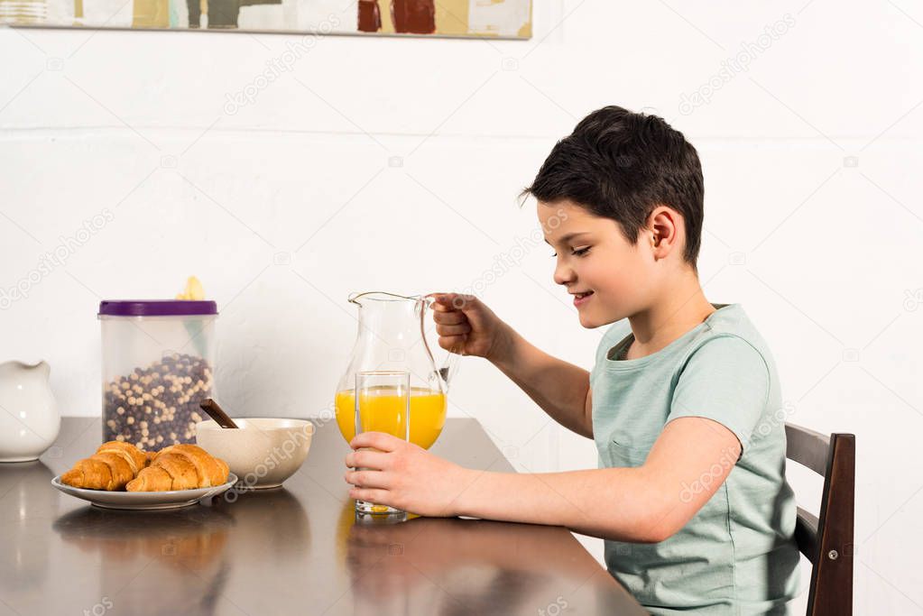 smiling boy holding glass and jug with orange juice in kitchen