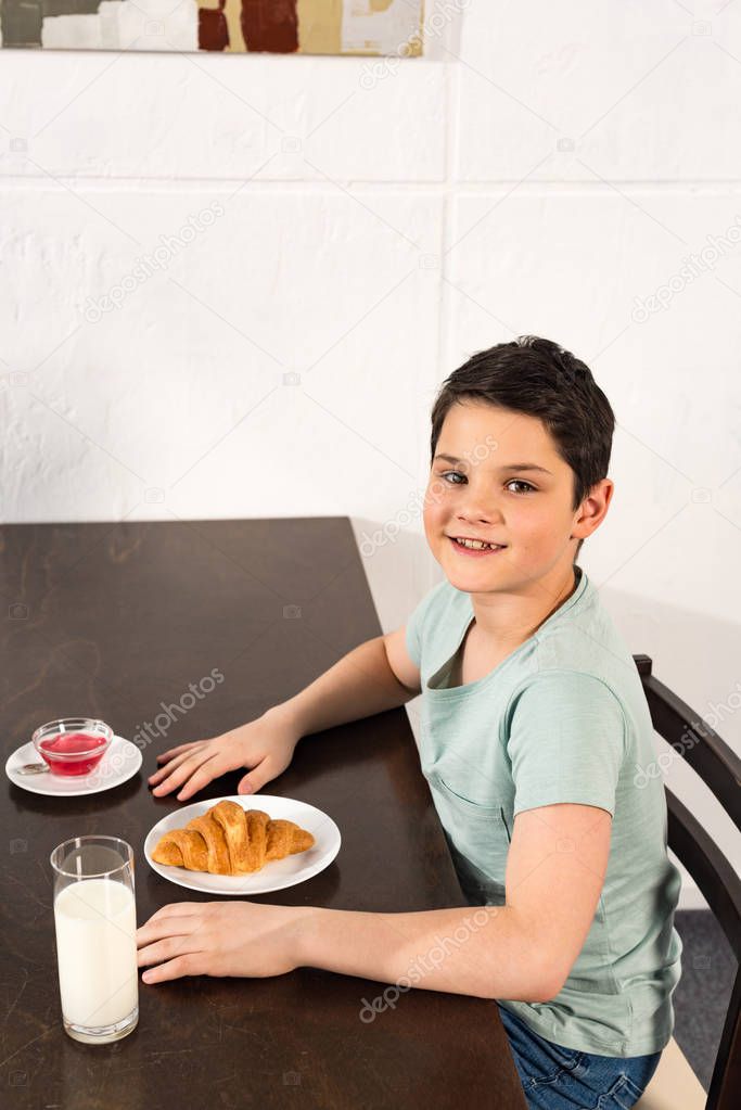 overhead view of smiling boy sitting at table with croissant, syrup and glass of milk