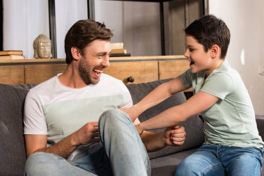 laughing son and dad sitting on sofa and having fun in living room clipart