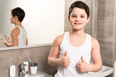 smiling boy standing near mirror in bathroom and showing thumbs up clipart