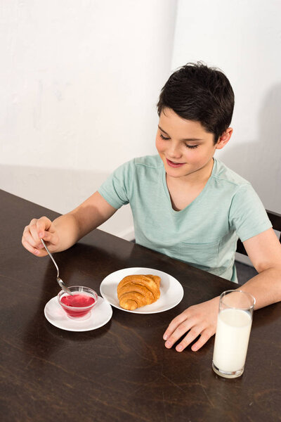 overhead view of smiling boy sitting at table with croissant, syrup and glass of milk