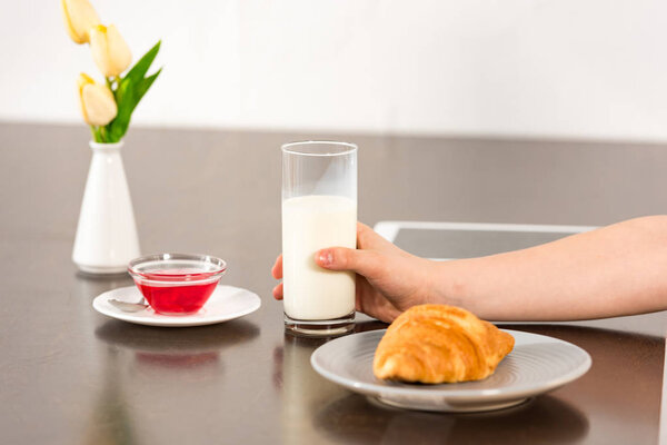cropped view of boy sitting at table with croissant, syrup and glass of milk