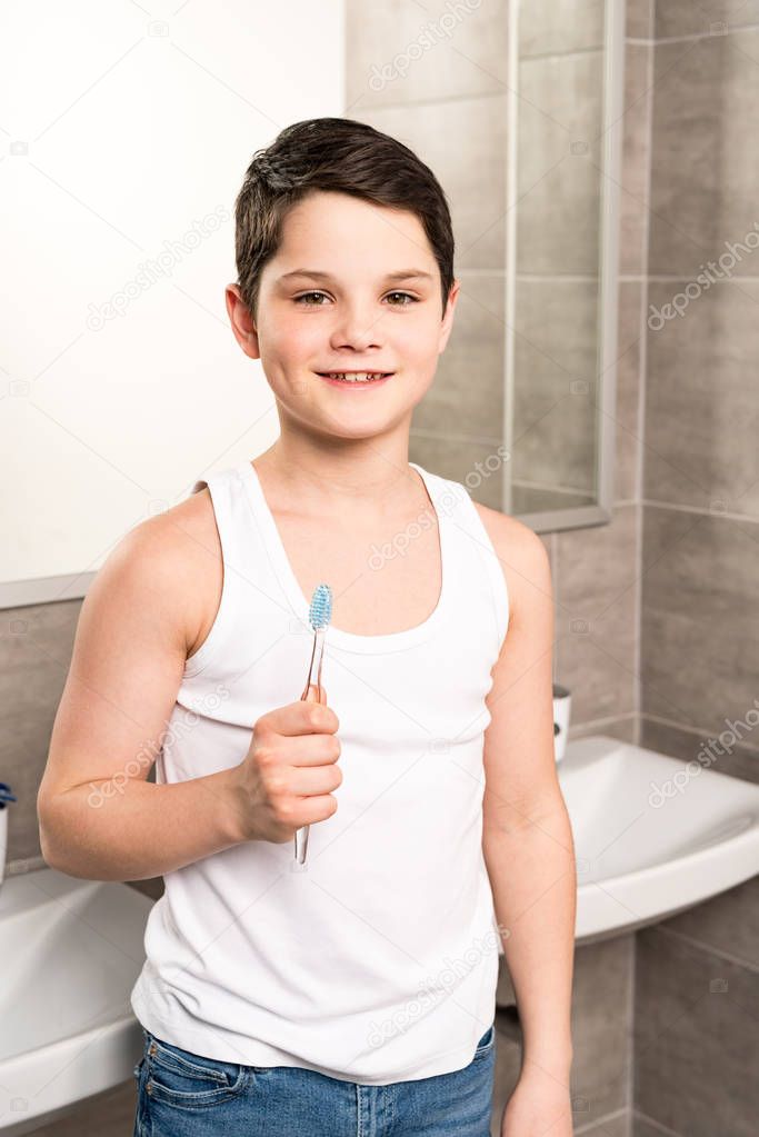 smiling boy holding toothbrush and looking at camera in bathroom