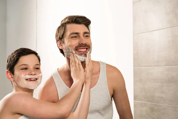 青春期前男孩在浴室里给微笑的父亲涂抹剃须泡沫 — 图库照片