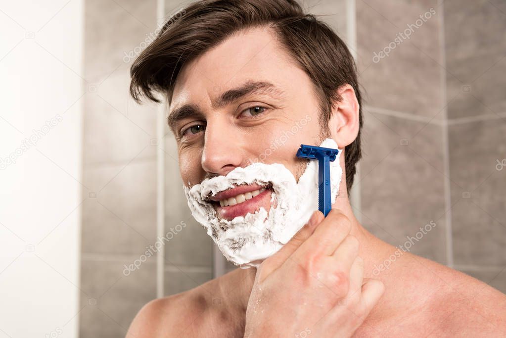 smiling man shaving beard with razor in bathroom