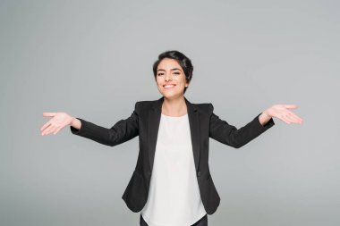 happy mixed race businesswoman showing welcoming gesture and smiling at camera isolated on grey clipart