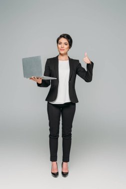 güzel karışık ırk iş kadını dizüstü bilgisayar tutan ve gri arka plan üzerinde başparmak gösteren