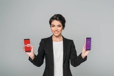 KYIV, UKRAINE - APRIL 24, 2019: Cheerful mixed race businesswoman holding smartphones with Youtube and Instagram apps on screen isolated on grey. clipart