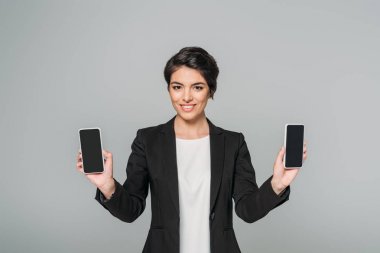 cheerful mixed race businesswoman showing smartphones with blank screen isolated on grey clipart