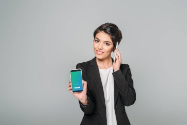 KYIV, UKRAINE - APRIL 24, 2019: Smiling mixed race businesswoman showing smartphone with Twitter app on screen while talking on smartphone isolated on grey. clipart