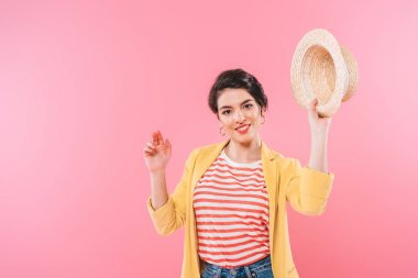 pretty mixed race woman waving straw hat and smiling at camera isolated on pink clipart