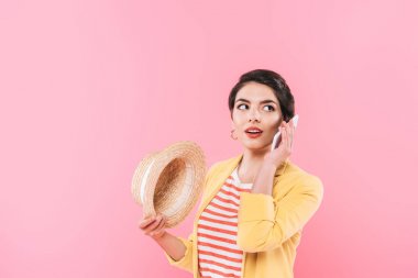 pretty mixed race woman talking on smartphone and holding straw hat isolated on pink clipart