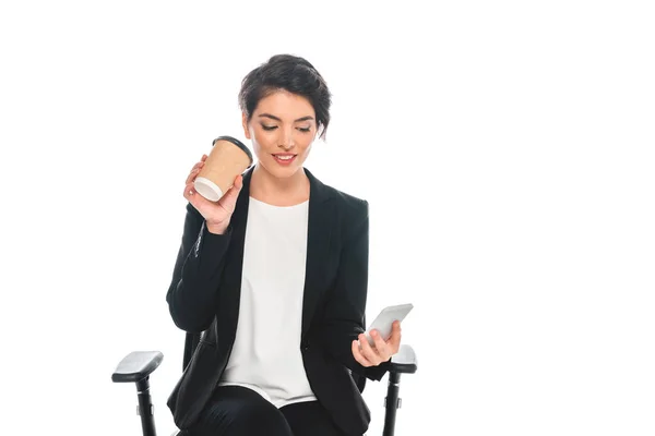 魅力的な混合レースビジネスウーマンは 紙コップからコーヒーを飲み 白で隔離されたオフィスチェアに座っている間 スマートフォンを使用しています — ストック写真