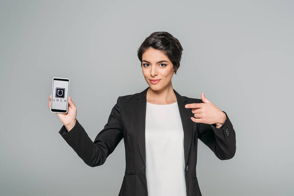 KYIV, UKRAINE - APRIL 24, 2019: Pretty mixed race businesswoman pointing with finger at smartphone with Uber app on screen isolated on grey.