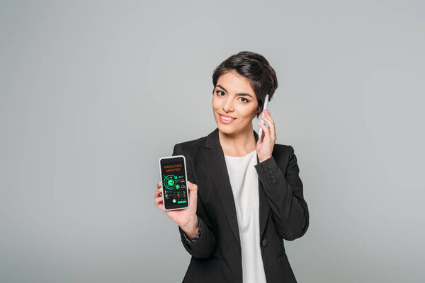 smiling mixed race businesswoman talking on smartphone and showing smartphone with marketing analysis app on screen isolated on grey