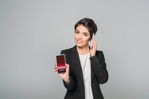 beautiful mixed race businesswoman showing smartphone with trading courses app on screen while talking on smartphone isolated on grey