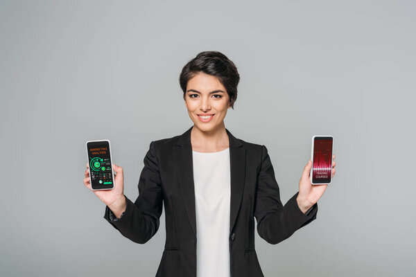smiling mixed race businesswoman holding smartphones with trading courses and marketing analysis apps on screen isolated on grey