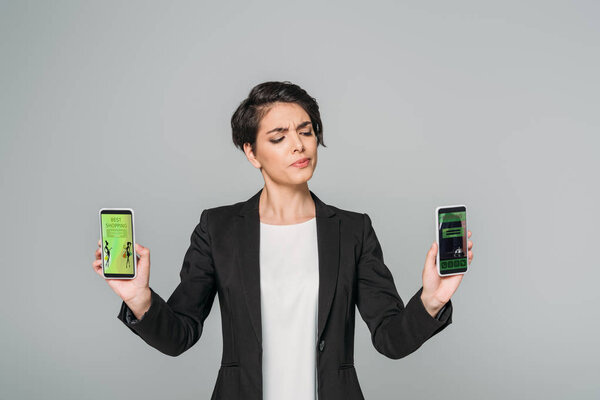 young mixed race skeptical businesswoman holding smartphones with best shopping and booking apps on screen isolated on grey
