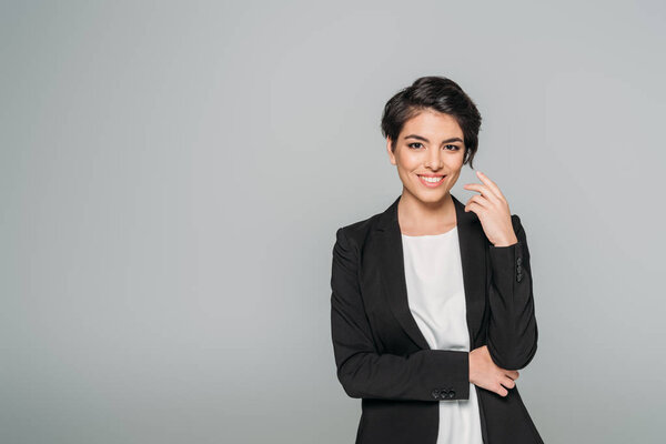 cheerful mixed race businesswoman in black formal wear smiling at camera isolated on grey