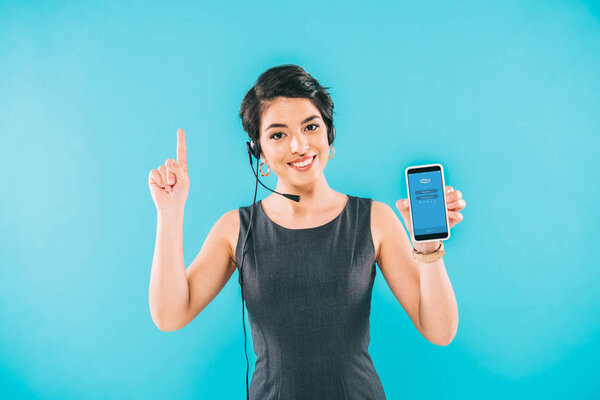 KYIV, UKRAINE - APRIL 24, 2019: Cheerful mixed race call center operator holding smartphone with Skype app on screen and showing idea sign isolated on blue.