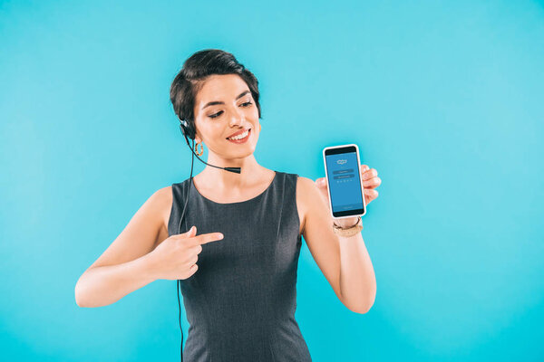 KYIV, UKRAINE - APRIL 24, 2019: Cheerful mixed race call center operator presenting smartphone with Skype app on screen isolated on blue.