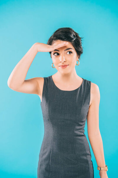 smiling mixed race woman holding hand near forehead and looking away isolated on blue