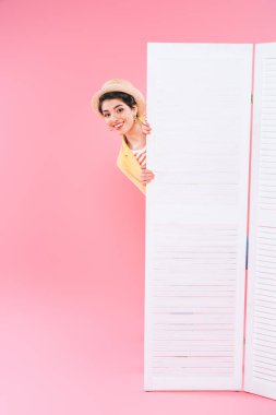 smiling mixed race woman looking out folding screen on pink background clipart