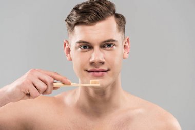 Gri üzerine izole edilmiş ahşap diş fırçasıyla dişlerini fırçalayan çıplak genç adamın ön görüntüsü
