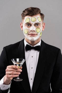 Resmi kıyafetli, salatalık maskeli genç bir adam. Elinde gri bir kokteyl bardağı var.
