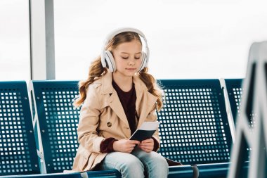preteen çocuk mavi koltuk üzerinde oturan, pasaport tutarak ve havaalanında bekleme salonunda müzik dinleme 