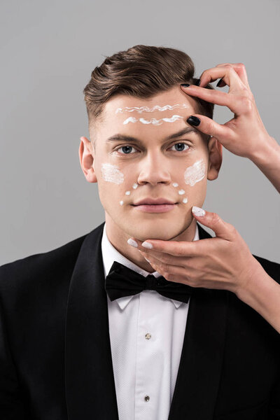 partial view of cosmetologists applying cream and man in formal wear isolated on grey