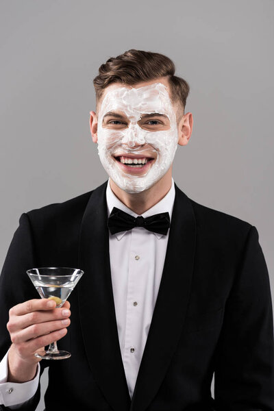 front view of smiling man in formal wear with cream on face holding glass of cocktail isolated on grey