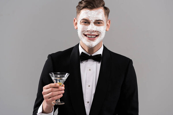 front view of smiling man in formal wear with cream on face holding glass of cocktail isolated on grey