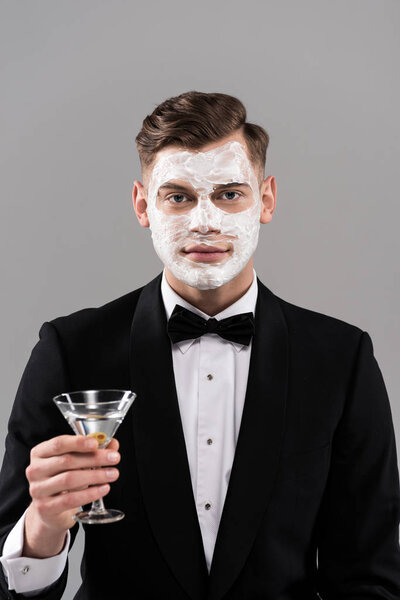 front view of man in formal wear with cream on face holding glass of cocktail isolated on grey