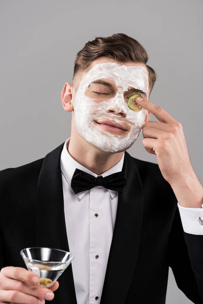 young man in formal wear with face mask holding glass of cocktail and applying cut cucumber on eye isolated on grey