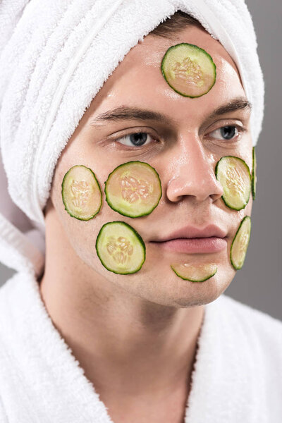 young man in bathrobe with cut cucumbers on face isolated on grey