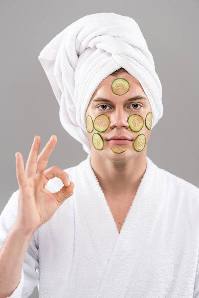 front view of young man in bathrobe with cut cucumbers on face showing okay sign isolated on grey