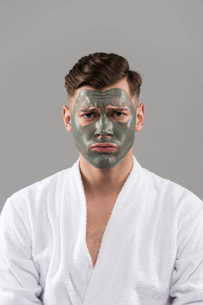 front view of sad young man in bathrobe with clay mask isolated on grey