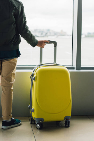 cropped view of boy holding yellow suitcase, standing near window in waiting hall