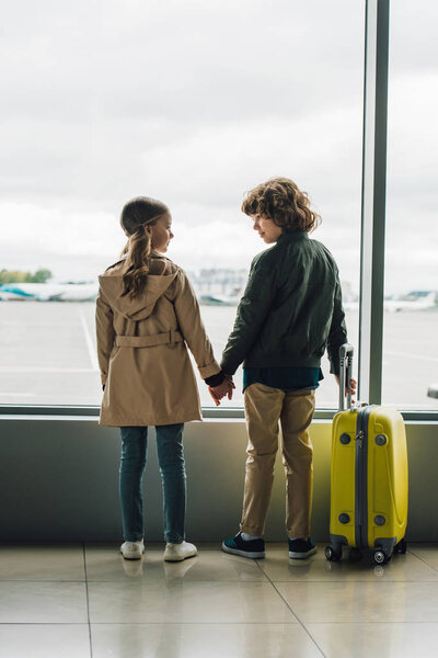 back view of kids holding hands and looking at each other near window in airport