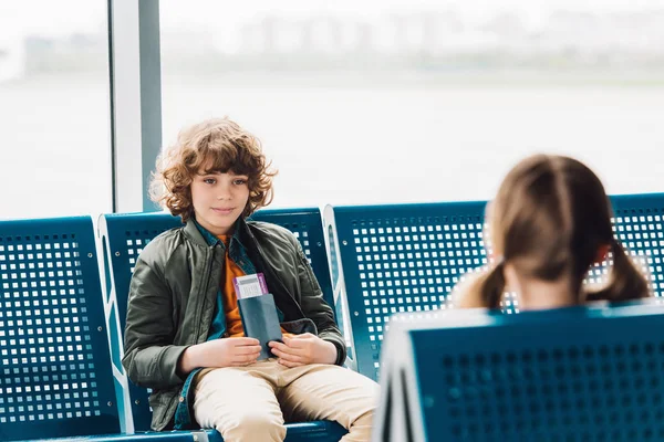 男孩坐在蓝色的座位上 拿着护照 看着在机场候机大厅的朋友 — 图库照片