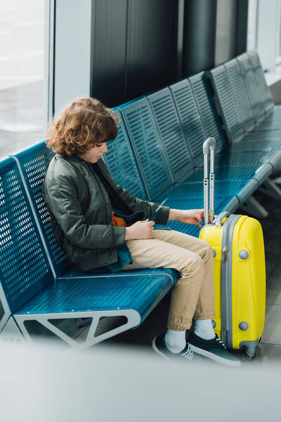 side view of boy holding passport and sitting on blue seat near yellow suitcase in waiting hall in airport 