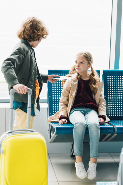 full length view of boy holding yellow suitcase and giving passport to preteen kid sitting on blue seat in waiting hall in airport 