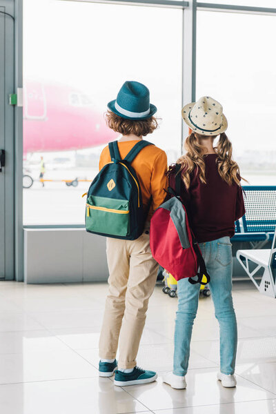 вид сзади на недоношенных детей с рюкзаками в зале ожидания
