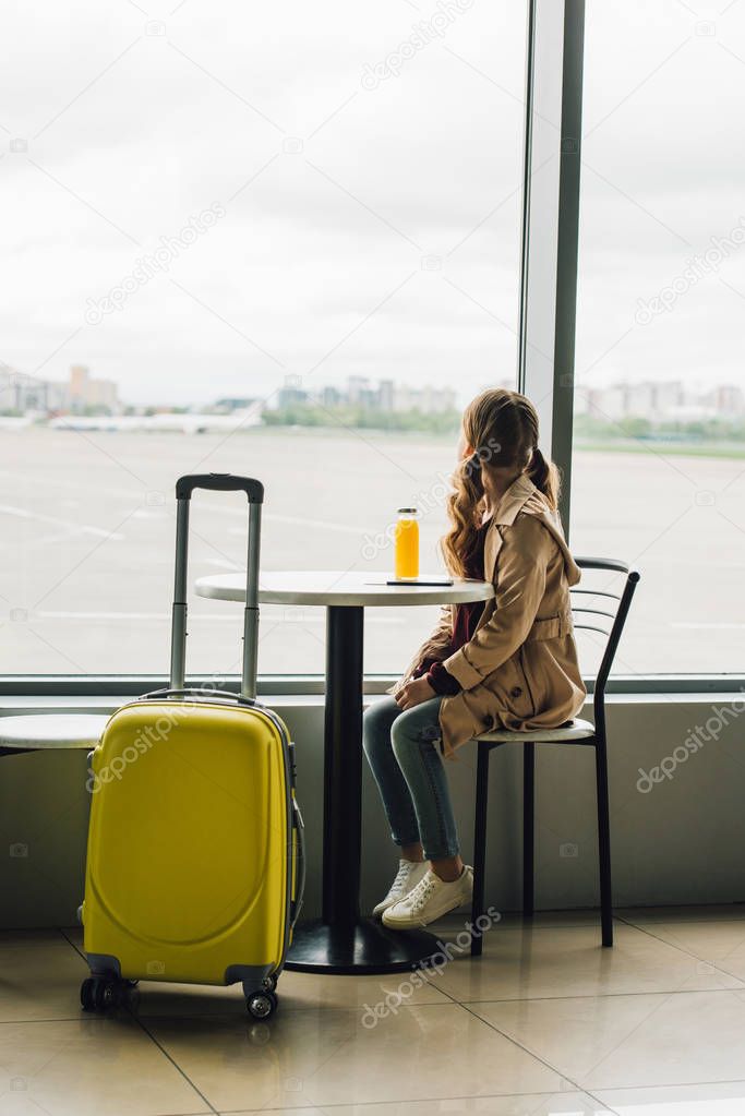 preteen kid sitting near yellow suitcase in waiting hall in airport and looking through window