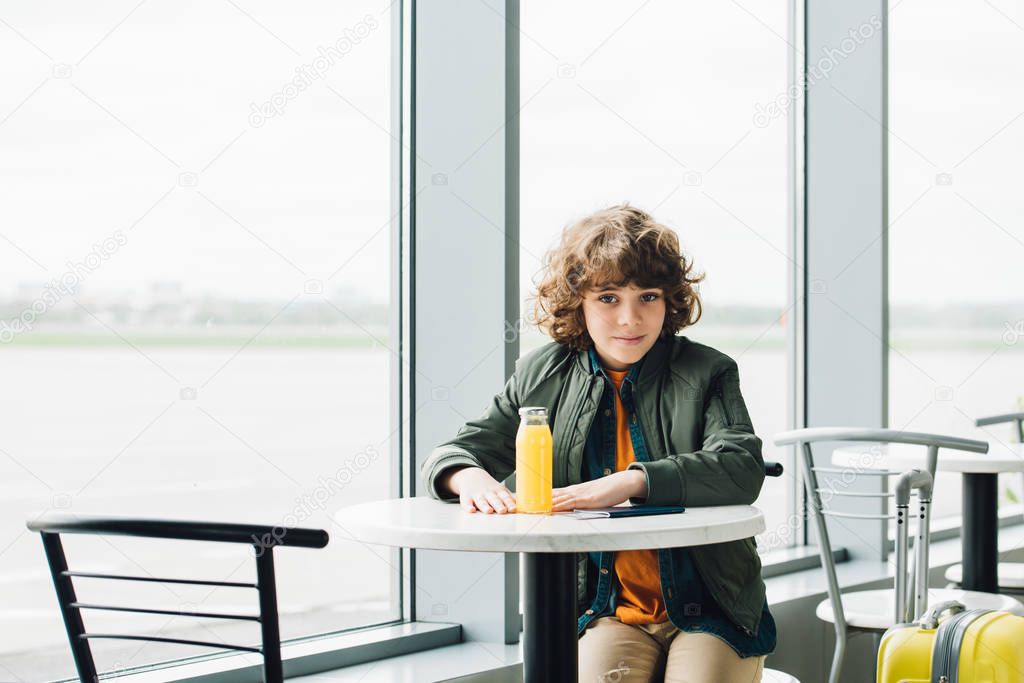boy sitting at table with orange juice on it and waiting hall in airport 