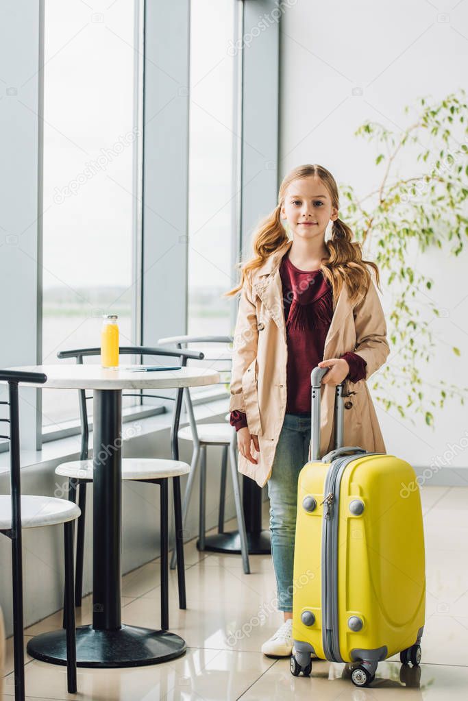 full length view of preteen kid standing near table and chair with yellow suitcase in airport