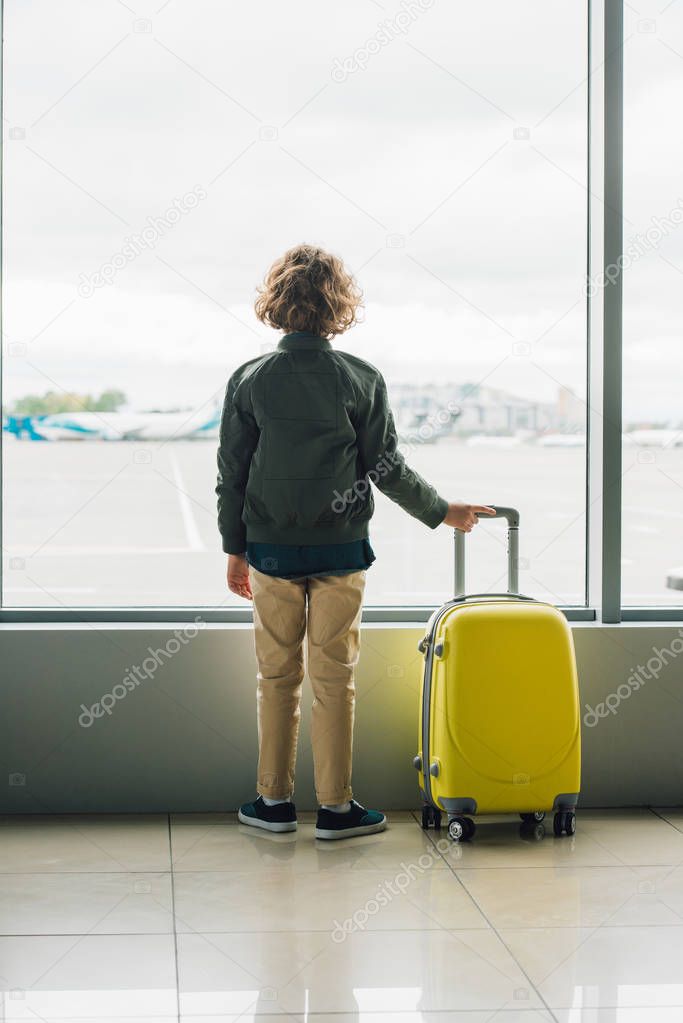 back view of boy standing near window, holding yellow suitcase